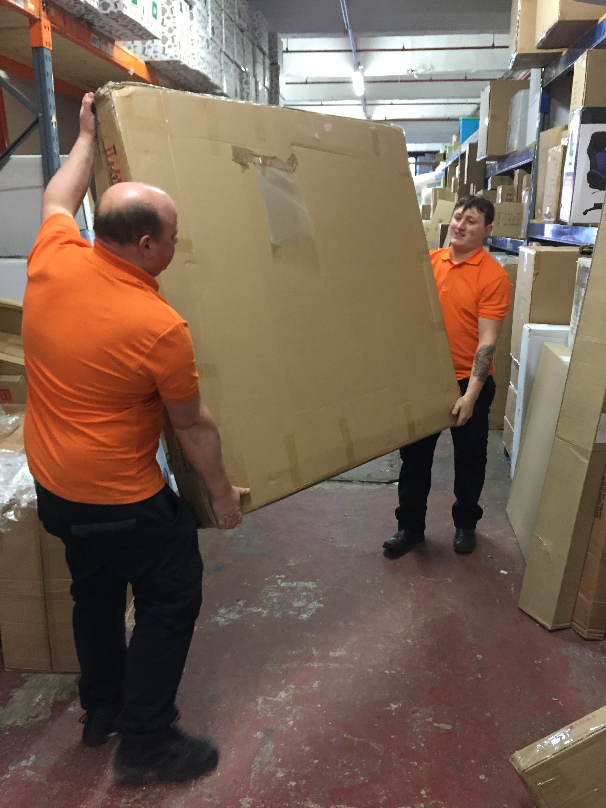 Two of the AJM Transport team in orange shirts carrying a large boxed item through a warehouse